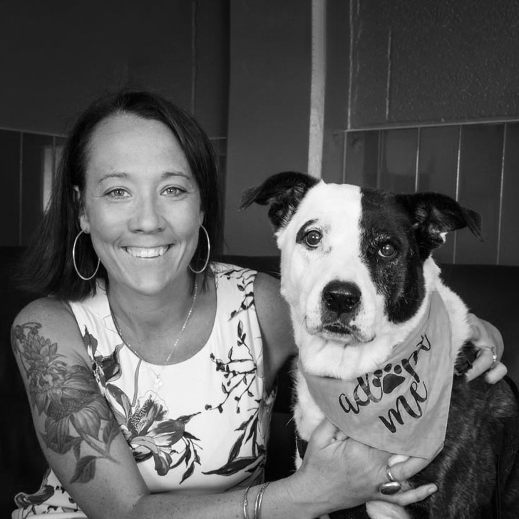 Kristen Hassen (Outcomes Consulting for animal welfare organizations) hugging a dog wearing a "adopt me" bandana, black and white photo.